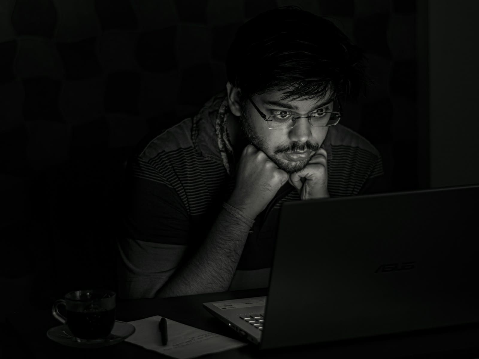 A person resting their chin on their hands, deep in thought, lit only by a laptop screen in a dark room